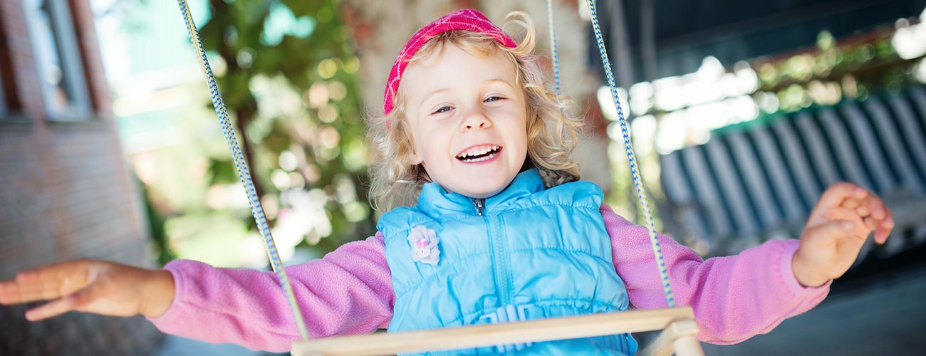 Bambina bionda con maglia rosa e gilet azzurro si dondola su un'altalena di legno